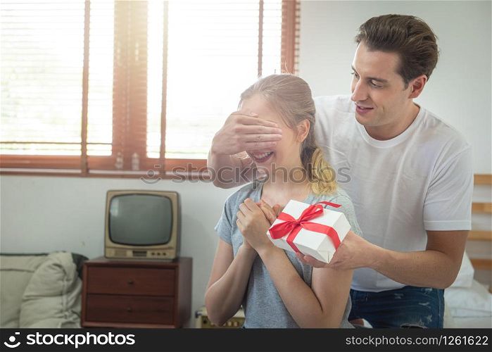 Young caucasian boyfriend surprising girlfriend with gift on bed.man covering eyes of girl and standing behind,birthday or congratulation or valentine day concept with copy space.