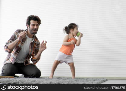 Young Caucasian father with beard and little daughter dancing together on the floor at home. Adorable child enjoy eating apple and swaying to music with dad