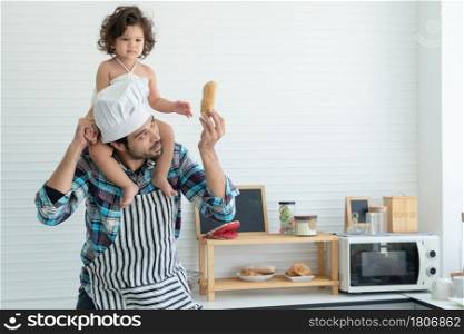 Young Caucasian father with beard wear chef hat and apron is giving homemade bread loaf to his little daughter kid who is riding neck in home kitchen with love and care. Fatherhood and child concept