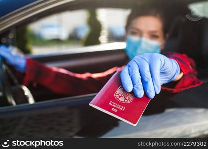 Young caucasian female driver wearing medical face mask and protective gloves handing over red immunity passport through car window,safety security border health check,Coronavirus COVID-19 pandemic
