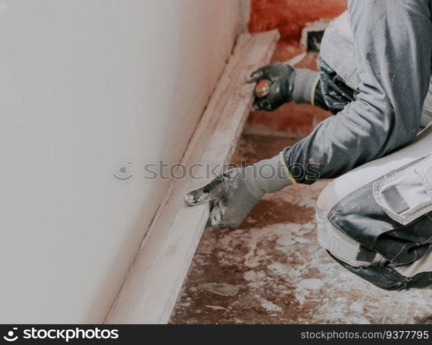 Young caucasian guy plasterer in gray long sleeve t-shirts, construction gloves holds a wooden rule and levels a freshly puttyed wall while squatting on the floor, close-up side view. The concept of apartment and house renovation, wall plastering, plastering services, at home.. Young caucasian guy plasterer holds a wooden rule and levels a freshly puttyed wall