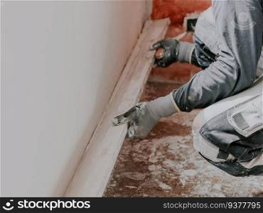 Young caucasian guy plasterer in gray long sleeve t-shirts, construction gloves holds a wooden rule and levels a freshly puttyed wall while squatting on the floor, close-up side view. The concept of apartment and house renovation, wall plastering, plastering services, at home.. Young caucasian guy plasterer holds a wooden rule and levels a freshly puttyed wall