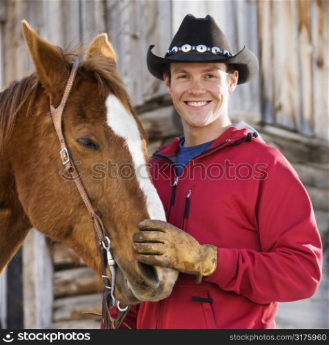 Young Caucasian man in cowboy hat petting his horse.