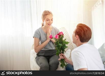 Young caucasian man sit down on knee gives bunch of roses to woman, happiness and surprise while they having a romantic date at home,valentine concept with copy space.