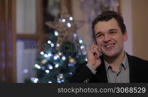 Young Caucasian man smiling while talking on the mobile phone, with a Christmas tree with white and blue lights in the background