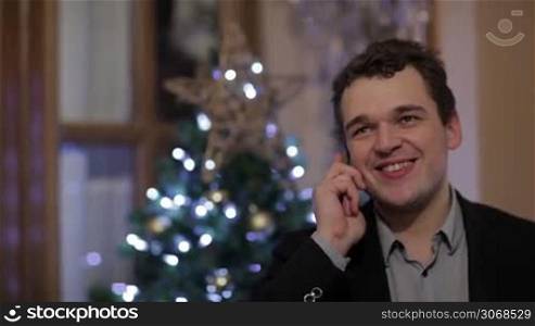 Young Caucasian man smiling while talking on the mobile phone, with a Christmas tree with white and blue lights in the background
