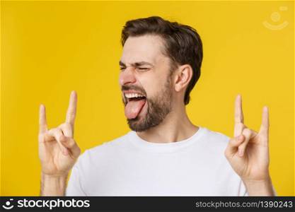 Young caucasian man wearing casual white t-shirt over yellow isolated background shouting with crazy expression doing rock symbol with hands up. Young caucasian man wearing casual white t-shirt over yellow isolated background shouting with crazy expression doing rock symbol with hands up.