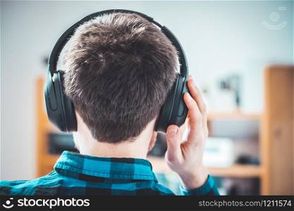 Young Caucasian man with checked blue shirt is listing to music with wireless headphones. Back of the head.