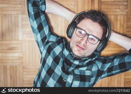 Young Caucasian man with checked blue shirt is lying on the wooden floor and listing to music with wireless headphones