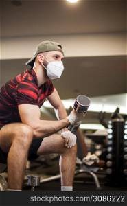 Young caucasian man working out wearing face mask & latex rubber gloves,performing bicep curl with dumbbells,COVID-19 pandemic social distancing rules while working out in indoor gym,United States US
