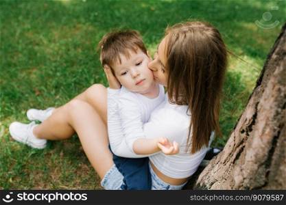 Young Caucasian mother kisses her three-year-old son on the cheek while sitting in the park near a tree on the grass in summer