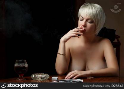 young Caucasian Nude blonde with bare Breasts sits at a table and smokes after losing at cards