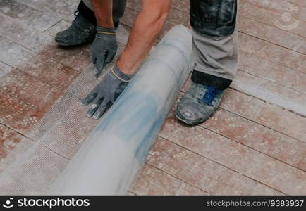 Young caucasian unrecognizable man in gray gloves cuts a construction mesh with a knife on a dirty wooden floor in a repair room, side view close-up top view with selective focus. The concept of home renovation, construction work.. One young man cuts construction mesh with a knife.