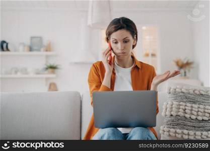 Young caucasian woman has laptop problem. Girl has consultation on phone. Lady is talking on telephone and telling some problem. Puzzled woman is sitting on sofa at home holding computer.. Young woman has laptop problem. Puzzled girl is sitting on sofa and telling talking on telephone.