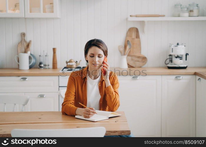 Young caucasian woman is call center assistant. Lady in orange shirt is remote worker at home. Girl is sitting at table at kitchen and taking notes into notebook. Customer support service.. Young woman is call center assistant. Girl is sitting at table at kitchen and taking notes.