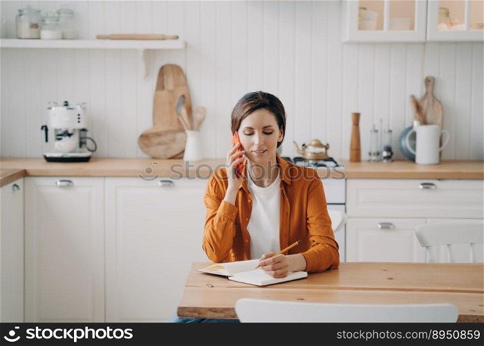 Young caucasian woman is call center assistant. Lady in orange shirt is remote worker at home. Girl is sitting at table at kitchen and taking notes into notebook. Customer support service.. Young woman is call center assistant. Girl is sitting at table at kitchen and taking notes.