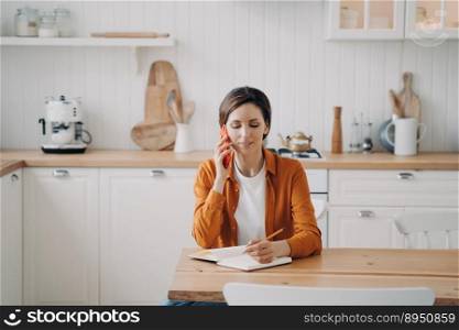 Young caucasian woman is call center assistant. Lady in orange shirt is remote worker at home. Girl is sitting at table at kitchen and taking notes into notebook. Customer support service.. Young woman is call center assistant. Girl is sitting at table at kitchen and taking notes.