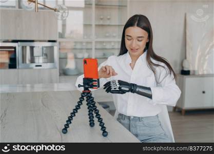 Young caucasian woman is filming blog with smartphone at home. Attractive handicapped girl is taking video how to use cyber arm. Beauty blogger has modern bionic prosthesis. Equality concept.. Young attractive handicapped beauty blogger is filming video with smartphone at home.