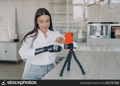 Young caucasian woman is filming blog with smartphone at home. Attractive handicapped girl is taking video how to use cyber arm. Beauty blogger has modern bionic prosthesis. Equality concept.. Young attractive handicapped beauty blogger is filming video with smartphone at home.