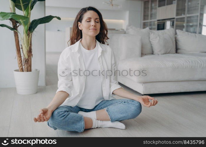 Young caucasian woman is sitting in lotus asana. Girl is practicing yoga and meditation on floor at home. Morning gymnastics. Harmony and mind relax concept. Scandinavian interior of living room.. Caucasian woman is sitting in lotus asana at home in morning. Scandinavian interior of living room.