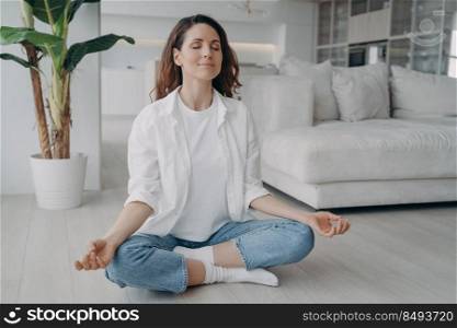 Young caucasian woman is sitting in lotus asana. Girl is practicing yoga and meditation on floor at home. Morning gymnastics. Harmony and mind relax concept. Scandinavian interior of living room.. Caucasian woman is sitting in lotus asana at home in morning. Scandinavian interior of living room.