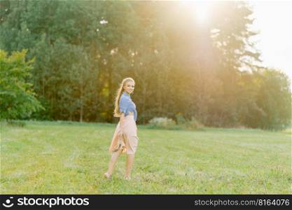 Young Caucasian woman or girl in a summer dress and a denim shirt is walking on the grass, holding shoes in her hands, relaxing outside the city on a weekend in summer