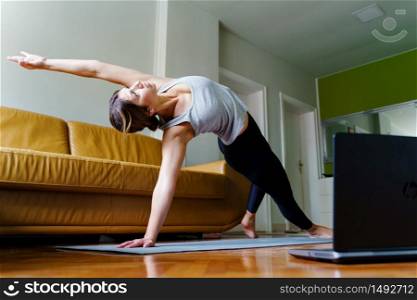 Young caucasian woman practicing yoga at home beautiful instructor teaching online class training in apartment standing in position asana in front of laptop using video in day or morning