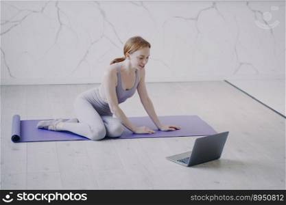 Young caucasian woman practicing yoga on mat on floor of her room. Internet fitness lesson with computer on self isolation. Concept of e-learning and home online classes on corona virus quarantine.. Young woman practicing yoga internet fitness lesson on self isolation. Home online classes.
