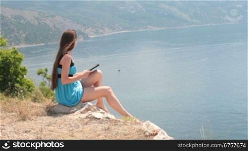 Young caucasian woman reads an ebook on a digital e-reader device on top of a mountain by the sea
