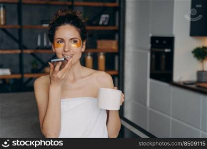 Young caucasian woman sending audio message. Girl applies anti wrinkle eye patches and relaxing at home talking on phone. Happy girl wrapped in towel after spa procedures. Body care and wellness.. Young caucasian woman sending audio message. Girl applies anti wrinkle eye patches relaxing at home.