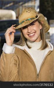 Young Caucasian woman tilting brim of her cowboy hat and smiling at viewer.