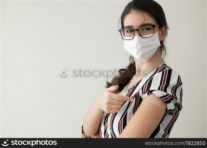 Young Caucasian woman wear face mask and glasses smile and pointing at plaster stuck on arm to show that she get vaccinated. Concept for people to get vaccinated to prevent corona virus. Copy space