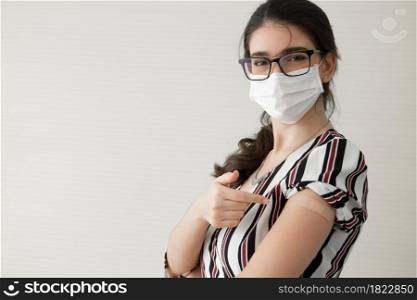 Young Caucasian woman wear face mask and glasses smile and pointing at plaster stuck on arm to show that she get vaccinated. Concept for people to get vaccinated to prevent corona virus. Copy space