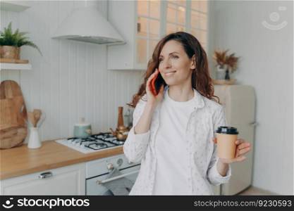 Young caucasian woman with phone has conversation in her stylish kitchen at home. Morning coffee at modern white scandinavian interior. Stove, worktop and cuisine.. Young caucasian woman with phone has coffee and conversation in her stylish kitchen at home.