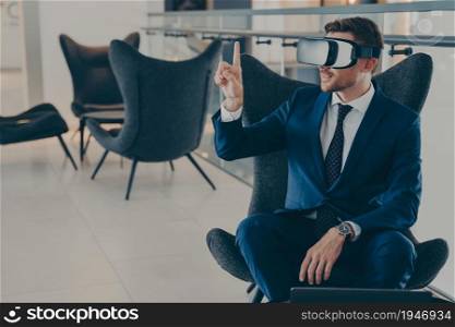 Young CEO sitting in office centre lobby with VR headset goggles , pointing with forefinger up in air, totally immersed in testing of virtual reality new business projects for company presentation. Young CEO sitting in office centre lobby with VR headset goggles, pointing with forefinger up in air