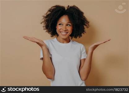 Young cheerful african woman can not make decision, spreading palms, dark skinned female with curly hair having no idea what happened while posing isolated over beige background. Hesitation and doubts. Young cheerful african woman spreading palms while posing isolated over beige background