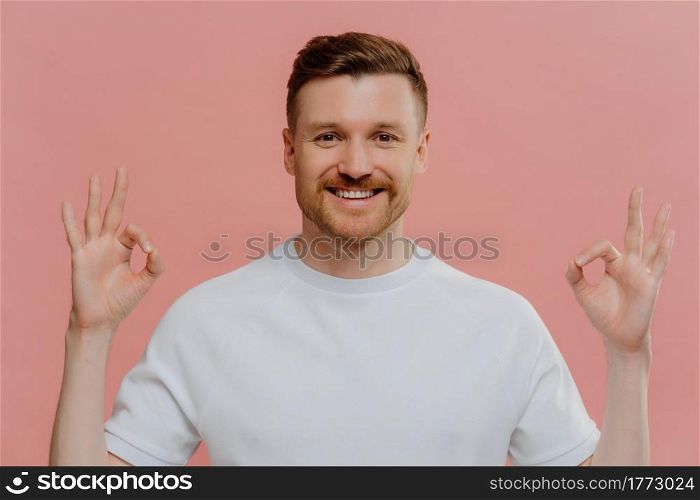 Young cheerful bearded man in white tshirt showing ok sign gesture with both hands, demonstrating agreement with positive face expression while standing against pink background. Body language concept. Young happy man showing okay gesture with both hands