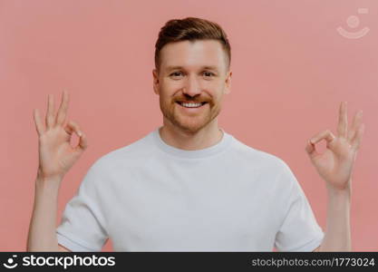 Young cheerful bearded man in white tshirt showing ok sign gesture with both hands, demonstrating agreement with positive face expression while standing against pink background. Body language concept. Young happy man showing okay gesture with both hands