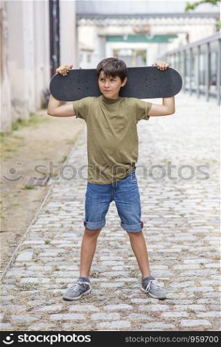 Young cheerful boy standing on the street while holding a skateboard while looking away in a bright day