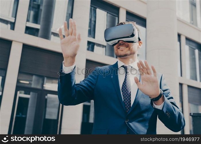 Young cheerful businessman with beard dressed in blue formal suit trying out VR glasses, checking out virtual reality, exploring digital world, standing alone next to building in background. Young businessman with beard dressed in blue formal suit trying out VR glasses outdoors