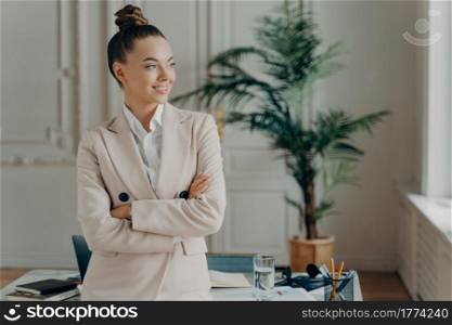 Young cheerful caucasian business woman dressed in formal elegant outfit posing near table in light office and looking aside with happy face expression, feeling satisfied with work done. Young female director in formal wear posing near table in light office