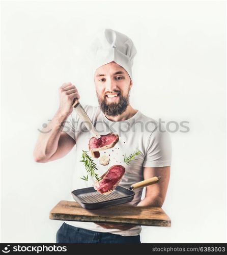 Young cheerful cook man with beard holding a frying pan with steaks and knife flying in the air on light background. Meat food and cooking concept