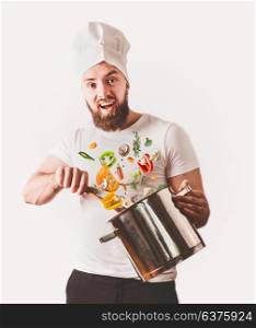 Young cheerful cook man with beard holding pan and spoon with flying vegetables on white background. Cooking concept