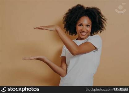 Young cheerful dark skinned showing large object, shaping box with both hands and smiling at camera while posing isolated over beige wall with copy space for text. Advertisement concept. Young dark skinned showing large object, shaping box with both hands, isolated over beige background