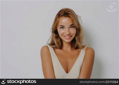 Young cheerful fair haired European woman with makeup smiles tenderly has healthy clean skin looks directly at camera dressed in casual wear poses in studio against grey background. Beauty concept. Young cheerful fair haired European woman with makeup smiles tenderly