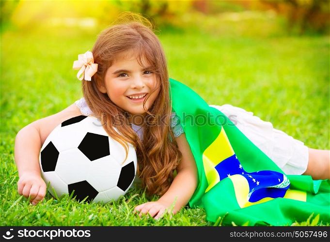 Young cheerful football fan lying down on fresh green grass with ball and big Brazil flag, happy supporter of Brazilian football team