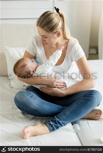 Young cheerful mother sitting on bed with her 3 months old baby boy