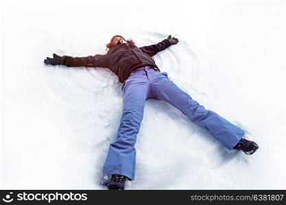 Young cheerful woman lying down on the white clean snow making snow angel, enjoying wintertime weather, happy winter holidays