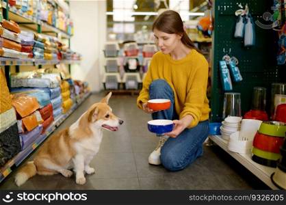 Young cheerful woman offering her lovely corgi dog to choose new feed bowl. Shopping at pet store. Young woman offering her lovely dog to choose new feed bowl