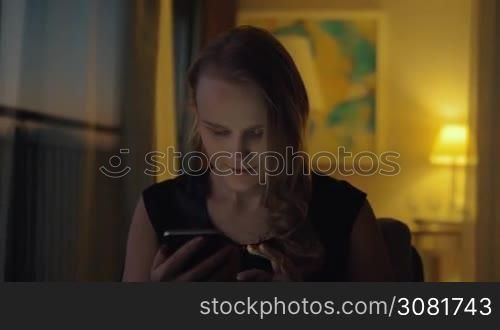 Young cheerful woman using smart phone at home in the evening. Shot in dim light of lamp
